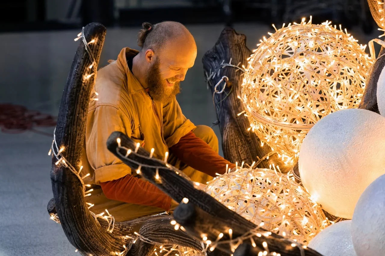Un artesano ensambla a mano, con una sonrisa y bajo una luz cálida, esferas luminosas meses antes de las fiestas