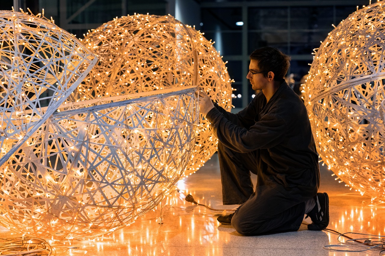 Un artesano ensambla a mano esferas luminosas de ratán y guirnalda cálida, de noche, en un taller vacío, mucho antes de diciembre